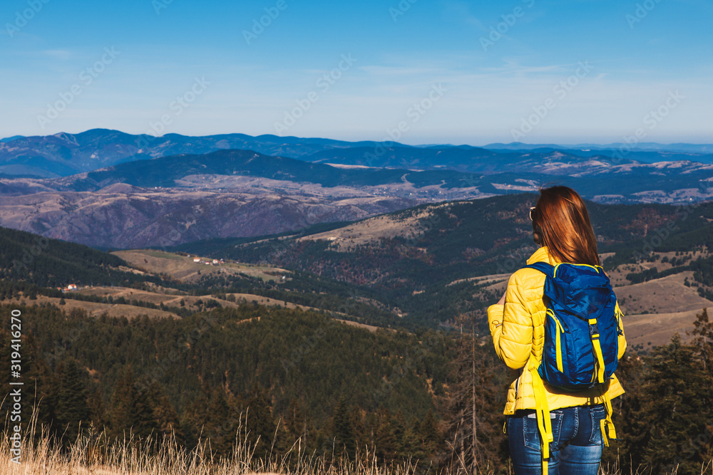 Rear view of female hiker with backpack standing on top of the mountain enjoying the view during the day.