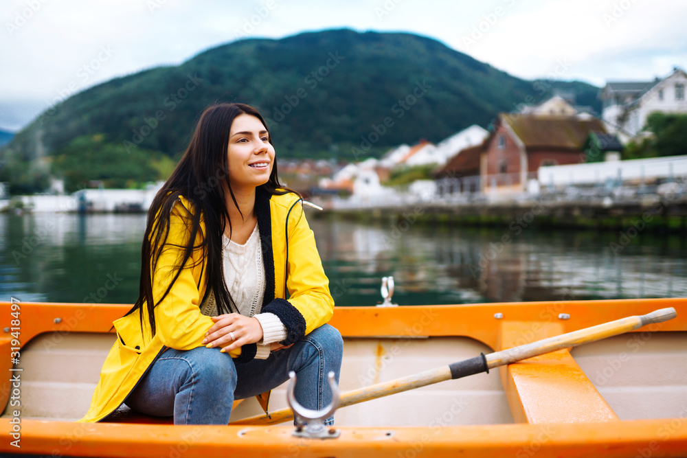 The girl tourist in a yellow jacket is sitting and posing in a boat against the backdrop of the mountains in the Norway. Young woman relaxing on the boat by the lake. Travelling, lifestyle, adventure. The girl tourist in a yellow jacket is sitting and posing in a boat against the backdrop of the mountains in the Norway. Young woman relaxing on the boat by the lake. Travelling, lifestyle, adventure.