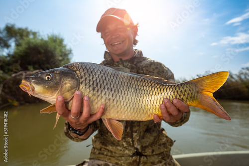 Murais de parede Happy angler holds big carp fish (Cyprinus carpio) and smiles