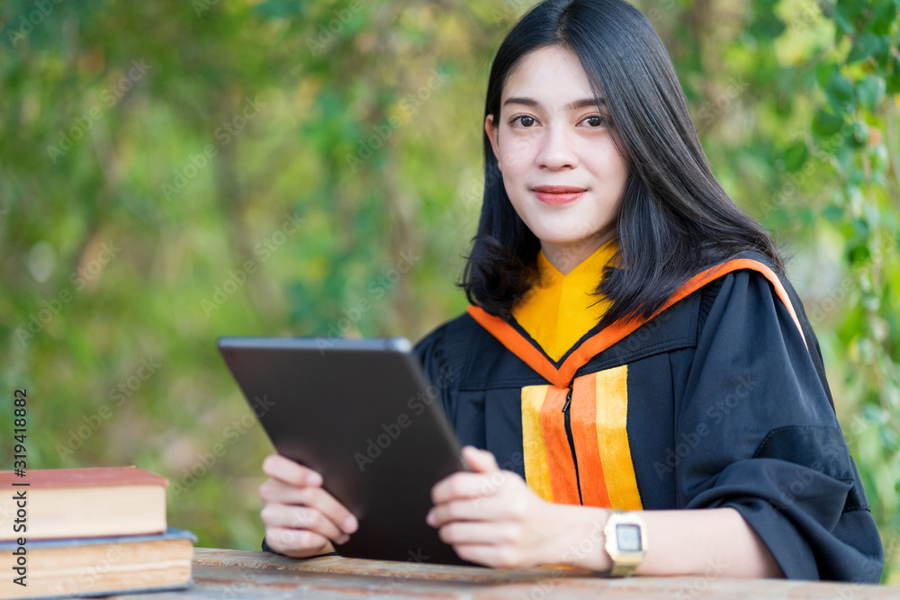 A young cheerful beautiful female gradute student wearing academic gown studying in college campus with books and lablet on table. A young cheerful beautiful female gradute student wearing academic gown studying in college campus with books and lablet on table.