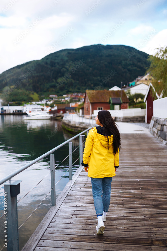 The girl tourist in a yellow jacket walking along the promenade in the Norway. Young woman smilling and posing against the backdrop of the mountains. Travelling, lifestyle, adventure concept.