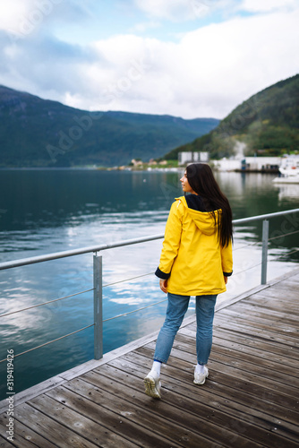 The girl tourist in a yellow jacket walking along the promenade in the Norway. Young woman smilling and posing against the backdrop of the mountains. Travelling, lifestyle, adventure concept.