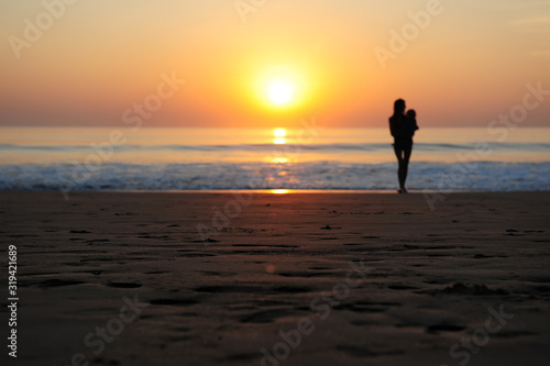 Silhouette of woman with a child in her arms on the background of sunset by the sea. Young slender girl carries a baby in the rays of setting sun. Beautiful mother walking along the sandy beach.