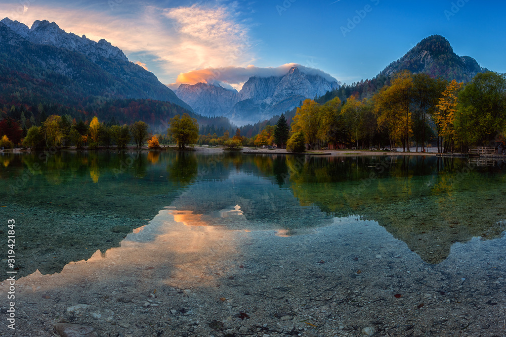 Jasna lake in Triglav national park at sunrise, Kranjska Gora, Slovenia. Amazing autumn landscape with Alps mountains, trees, blue sky with clouds and reflection in water, famous tourist attraction