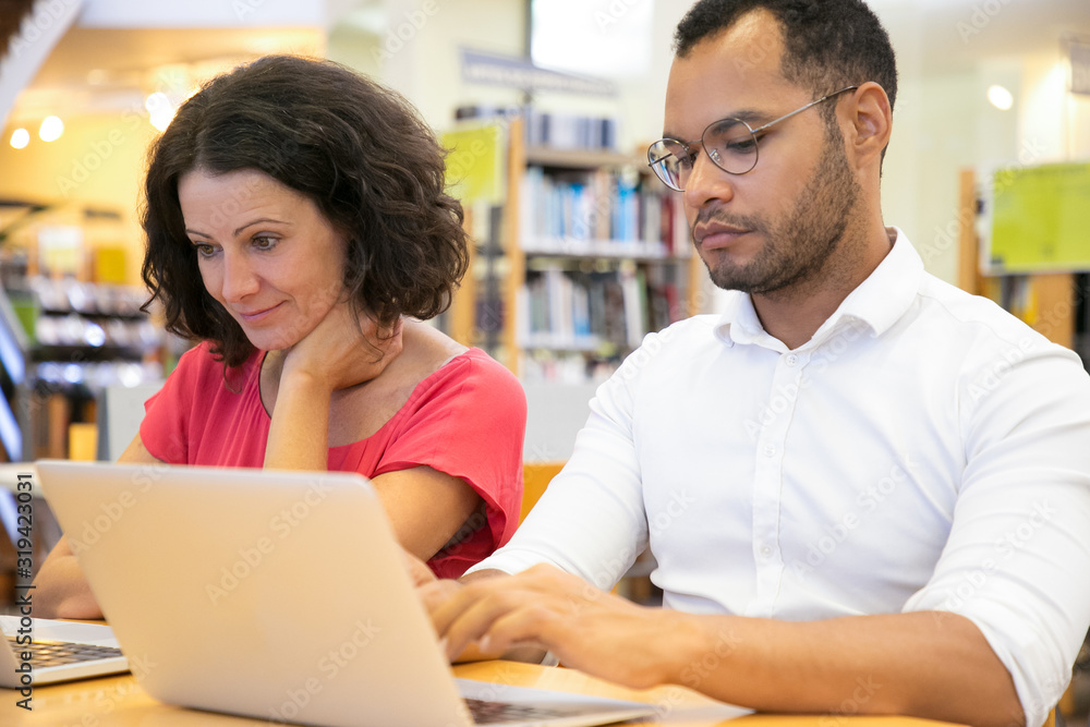 Two students using laptops while sitting at library. Smiling Caucasian woman and focused African American man studying with laptops together. Technology, education concept Two students using laptops while sitting at library. Smiling Caucasian woman and focused African American man studying with laptops together. Technology, education concept