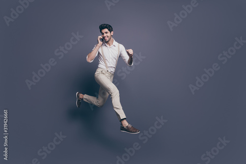 Full body profile side photo of excited guy speak smart phone listen discounts win lottery jump run wear white shirt pants trousers isolated over grey color background