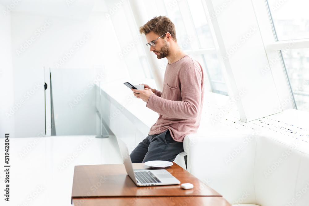 Fototapeta premium Image of young man using smartphone and working on laptop in cafe