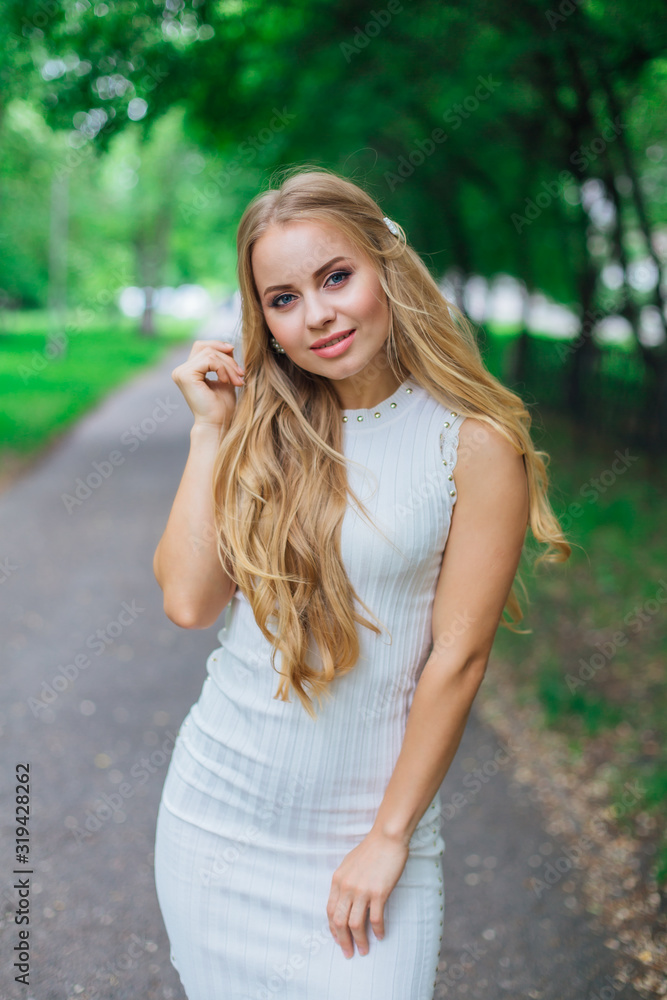 Portrait of a charming blond woman wearing beautiful white dress standing on the road under trees. Portrait of a charming blond woman wearing beautiful white dress standing on the road under trees.