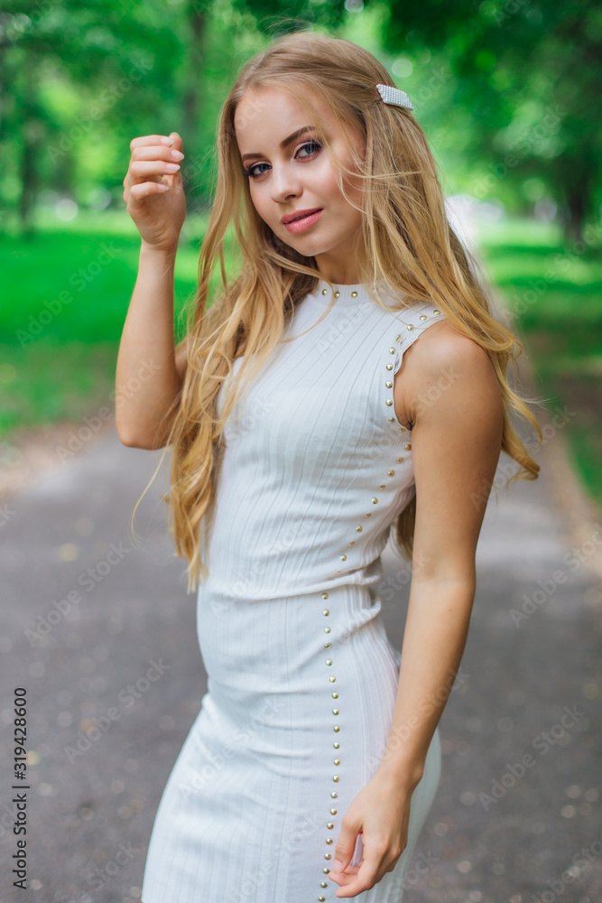 Portrait of a charming blond woman wearing beautiful white dress standing on the road under trees. Portrait of a charming blond woman wearing beautiful white dress standing on the road under trees.