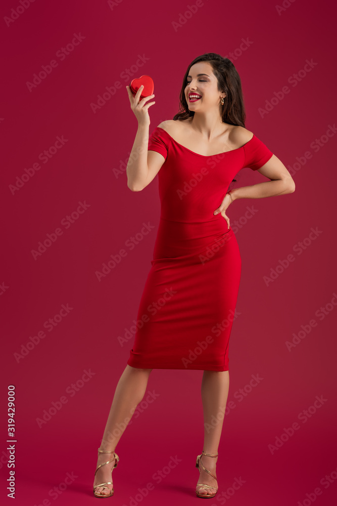 sensual, elegant girl holding heart-shaped gift box while standing with hand on hip on red background sensual, elegant girl holding heart-shaped gift box while standing with hand on hip on red background