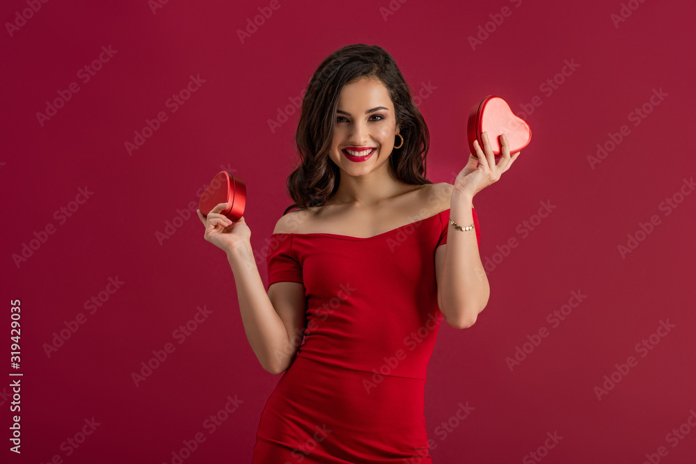 seductive, elegant girl holding heart-shaped gift boxes and smiling at camera isolated on red seductive, elegant girl holding heart-shaped gift boxes and smiling at camera isolated on red