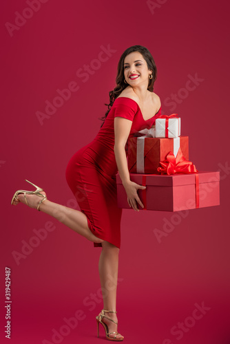 flirty, elegant girl holding gift boxes and looking away while standing on one leg on red background © LIGHTFIELD STUDIOS