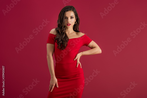 seductive girl in elegant dress looking at camera while posing with hand on hip isolated on red