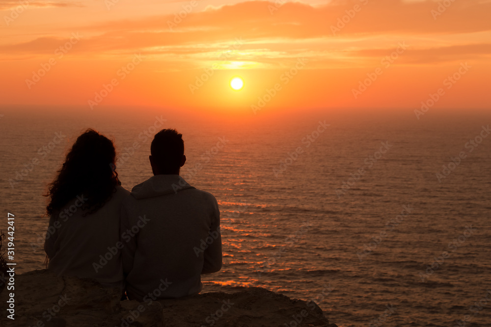 A couple sitting on a rock watching the sunset in front of the sea
