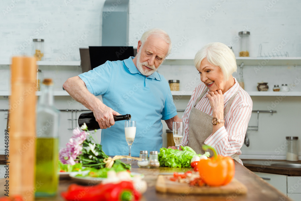 Selective focus of smiling woman looking at husband pouring champagne by bouquet and vegetables on kitchen table Selective focus of smiling woman looking at husband pouring champagne by bouquet and vegetables on kitchen table