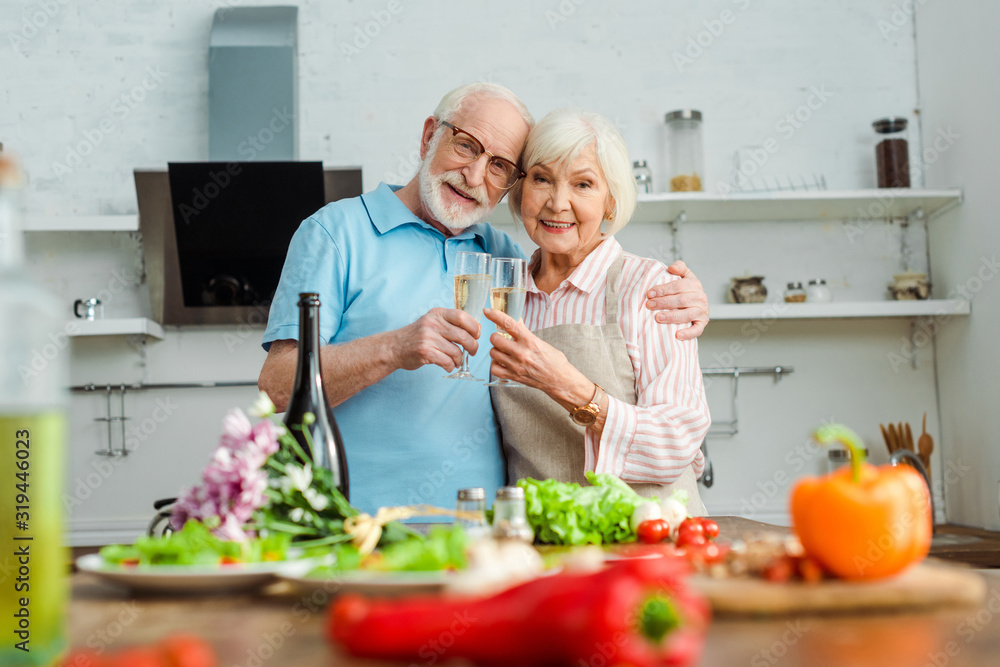 Selective focus of elderly couple smiling at camera while clinking with champagne by vegetables and bouquet on kitchen table Selective focus of elderly couple smiling at camera while clinking with champagne by vegetables and bouquet on kitchen table