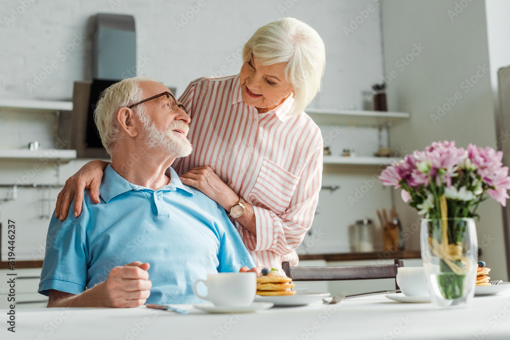 Selective focus of senior woman hugging smiling husband by coffee and pancakes on table Selective focus of senior woman hugging smiling husband by coffee and pancakes on table