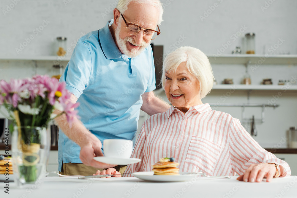 Selective focus of senior man putting coffee on table by smiling wife during breakfast Selective focus of senior man putting coffee on table by smiling wife during breakfast