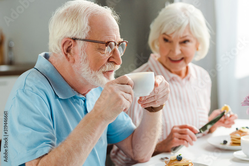 Selective focus of senior man drinking coffee by smiling wife with pancakes during breakfast