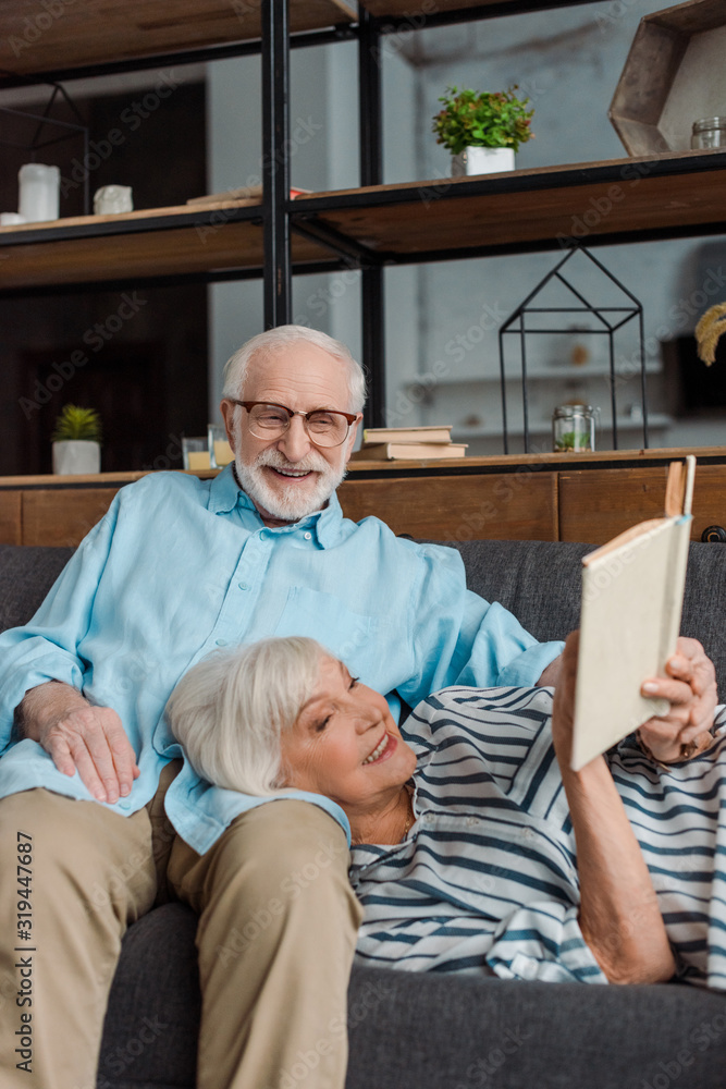 Senior woman reading book by smiling husband on couch at home Senior woman reading book by smiling husband on couch at home
