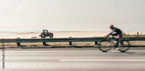 Competitive Professional Cyclist riding bicycle on Pacific Coast Highway in Santa Monica  California with a red tractor driving on the beach in the sand