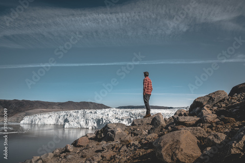 A young man traveler tourist standing in front of Eqip Sermia glacier called Eqi Glacier. Wall of ice in background. The concept of global warming and professional guides. On a sunny day with blue sky photo