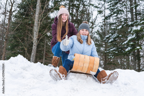 Obraz na plátně Two happy teen girls/friends sitting on a wood toboggan/sled while at the edge of a snow covered hill in a park and one girl holding her thumb up
