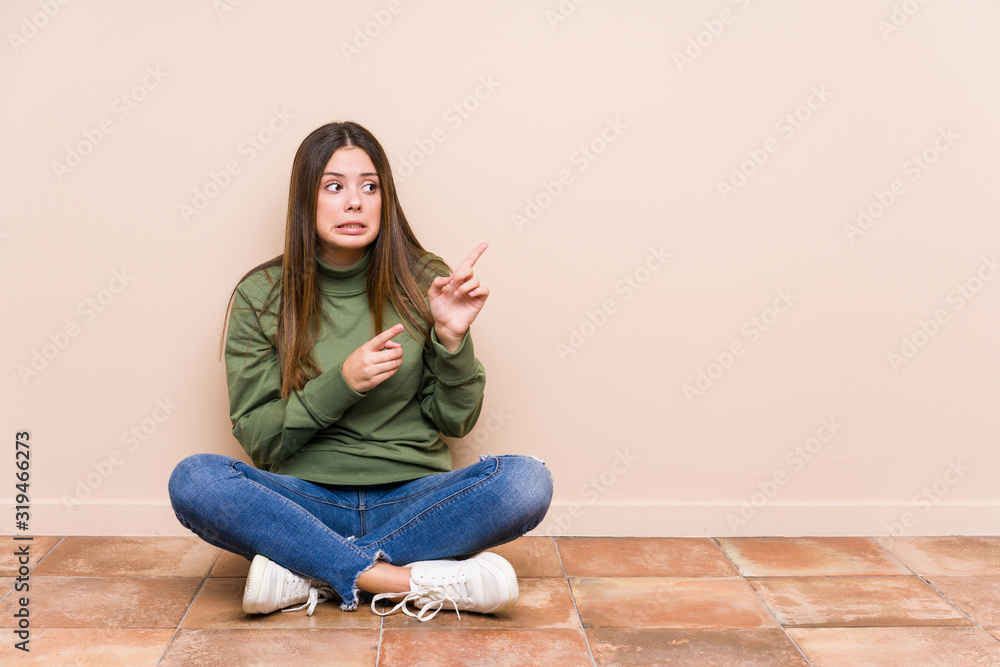 Young caucasian woman sitting on the floor isolated shocked pointing with index fingers to a copy space.