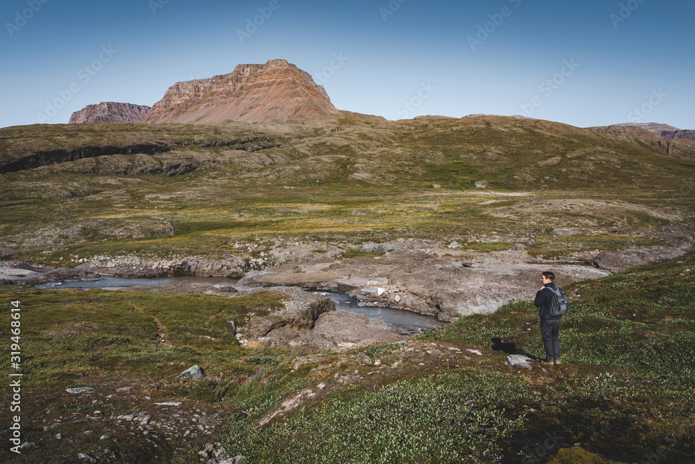 Young man hiker in the arctic landscape of Disko Bay in Greeland in Summer. Blue Sky and green meadows. Arctic Circle Trail with temple mountains. Disko Island and Village of Qeqertarsuaq,
