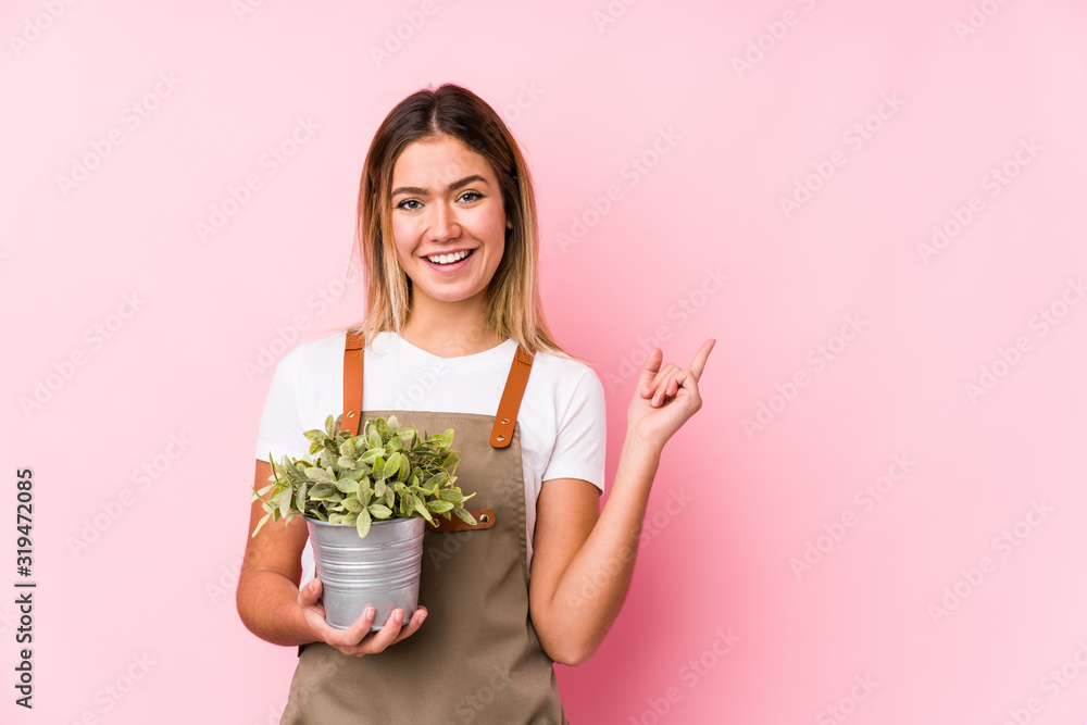 Young caucasian gardener woman in a pink background smiling and pointing aside, showing something at blank space. Young caucasian gardener woman in a pink background smiling and pointing aside, showing something at blank space.