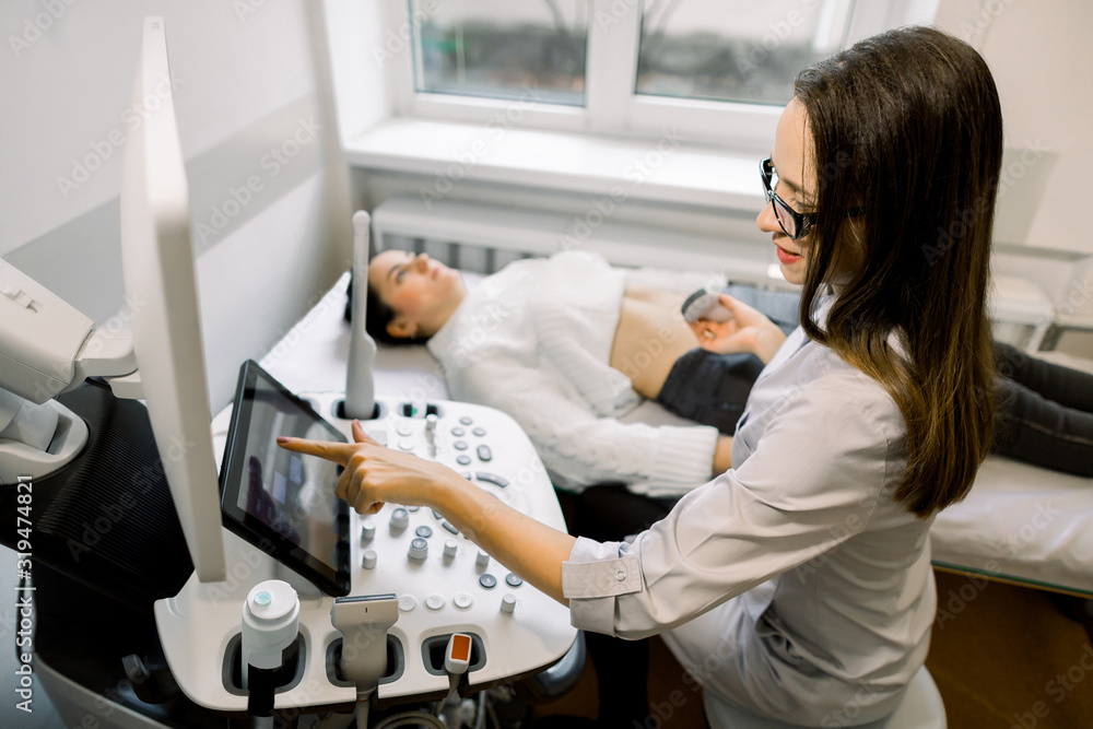 Obraz premium Young woman doctor preparing to the abdominal ultrasound for female patient. Doctor pushes buttons on a control panel before starting ultrasound sonogram procedure.