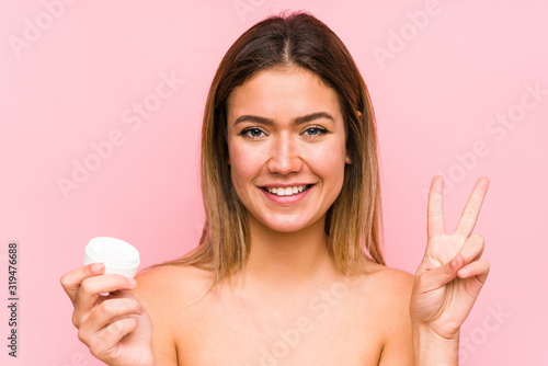 Young caucasian woman holding a moisturizer isolated showing number two with fingers.