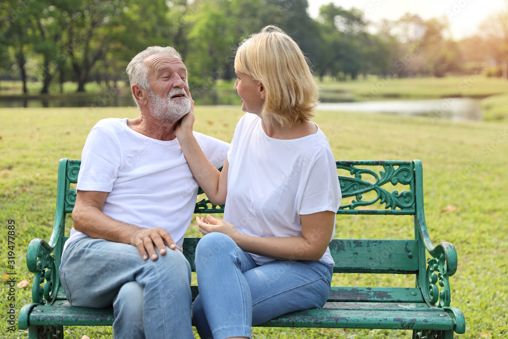 elderly caucasian couple with white shirt and blue jean sitting and embracing in park during summer time on wedding anniversary day