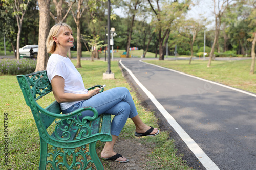 Senior caucasian woman in white T shirt and blue jeans with mobile phone sitting in the park outdoor during summer time. Elderly female looking away with green trees background. Lifestyle concept photo