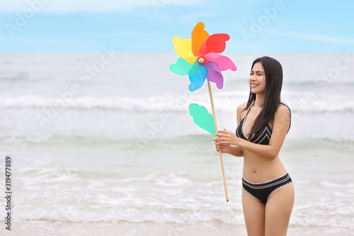 Young asian beautiful and sexy girl in black bikini who standing and holding rainbow colored windmills toy and smiling with happy and beauty face with blue sky on holiday vacation (lifestyle concept)