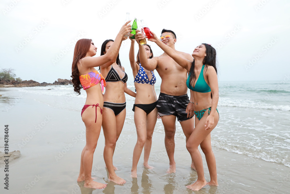 Group of happy asian man and women wearing bikini or swimming suit on the beach. They toasting beer bottles and soft drink bottle, having fun and happy time together. They are good body shape smiling. Group of happy asian man and women wearing bikini or swimming suit on the beach. They toasting beer bottles and soft drink bottle, having fun and happy time together. They are good body shape smiling.