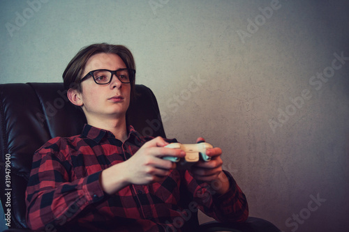 Serious adolescent boy playing video games late night seated relaxed in his armchair over grey wall background. Intent teen guy holding a joystick console looking attentive trying to win. photo