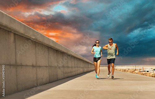 fitness  sport and healthy lifestyle concept - happy couple in sports clothes and sunglasses running along pier over sunset sky background
