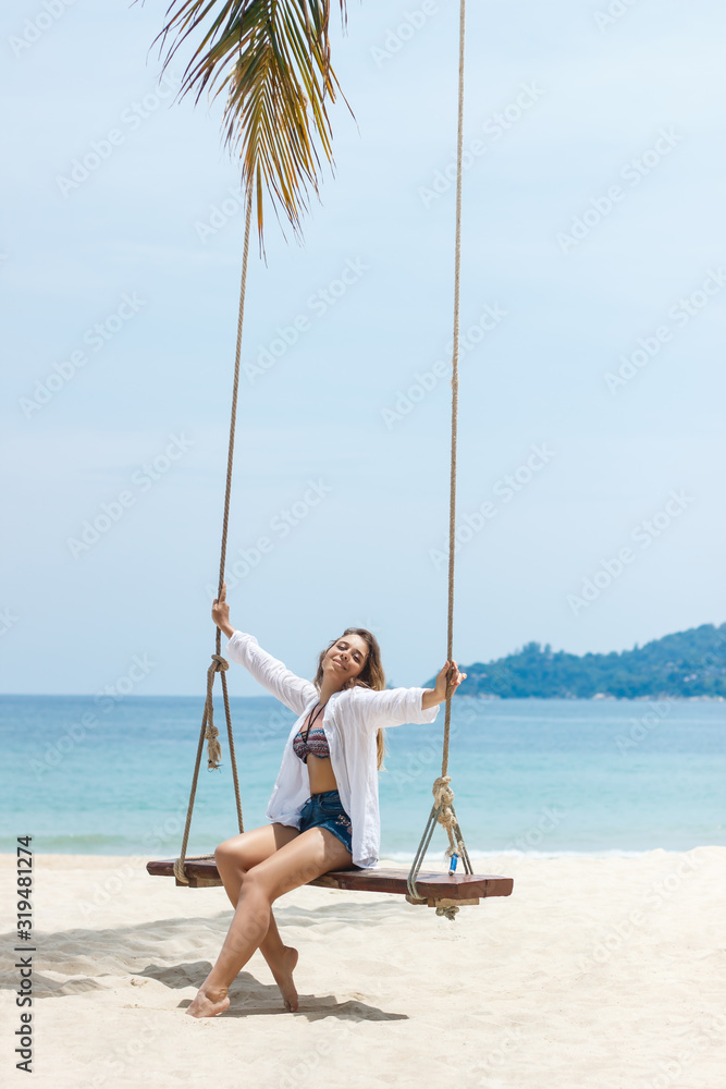 Tanned beautiful caucasian blonde young woman with a plumeria flower in her hair is swinging on a wooden swing on the shore of the blue sea. Sunny day, white sand. Wonderful paradise on earth