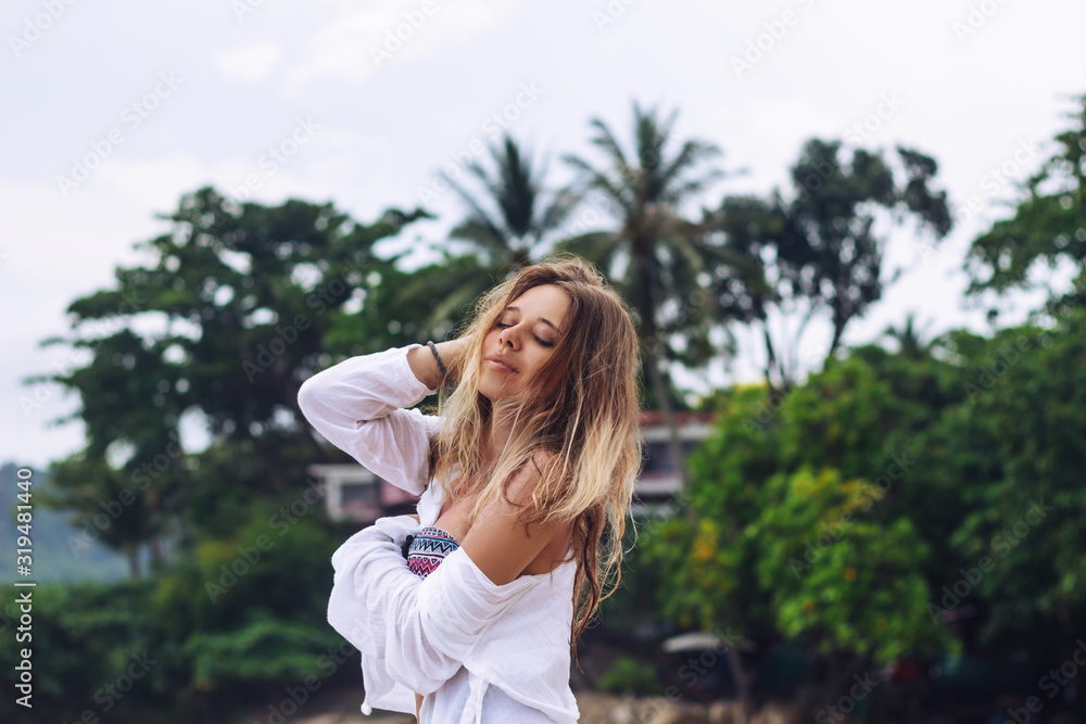 Tanned caucasian blonde young woman in a white shirt is enjoying the sun on the background of a palm trees. Paradise on earth Tanned caucasian blonde young woman in a white shirt is enjoying the sun on the background of a palm trees. Paradise on earth