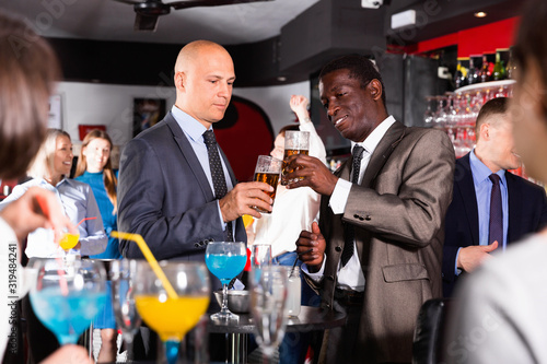 Two cheerful male colleagues enjoying corporate bar party, drinking beer and having fun conversation
