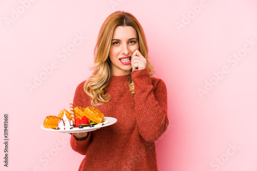 Young blonde woman eating a waffle dessert isolated biting fingernails, nervous and very anxious.