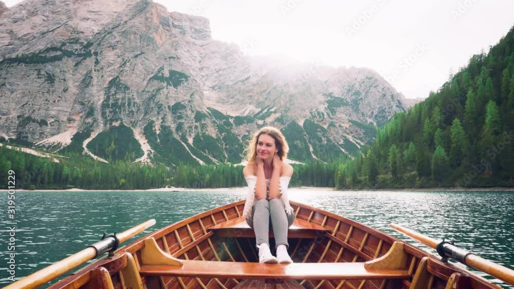 cute young girl sitting on edge wooden boat in vintage white boho shirt patterns bare shoulders, gray jeans. light picture mountains and azure lake turquoise water, beauty bright green nature Italy cute young girl sitting on edge wooden boat in vintage white boho shirt patterns bare shoulders, gray jeans. light picture mountains and azure lake turquoise water, beauty bright green nature Italy