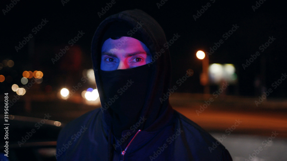 Man bandit in balaclava and hood looking at the camera at night blinking headlights colorful danger dangerous disguise gangster mask robber hooligan portrait terrorist unrecognizable white bad mugger Man bandit in balaclava and hood looking at the camera at night blinking headlights colorful danger dangerous disguise gangster mask robber hooligan portrait terrorist unrecognizable white bad mugger