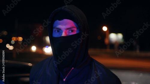 Man bandit in balaclava and hood looking at the camera at night blinking headlights colorful danger dangerous disguise gangster mask robber hooligan portrait terrorist unrecognizable white bad mugger