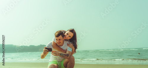 Happy couple taking selfies near sea. Loving couple embracing during date on beach against waving sea and cloudless sky.