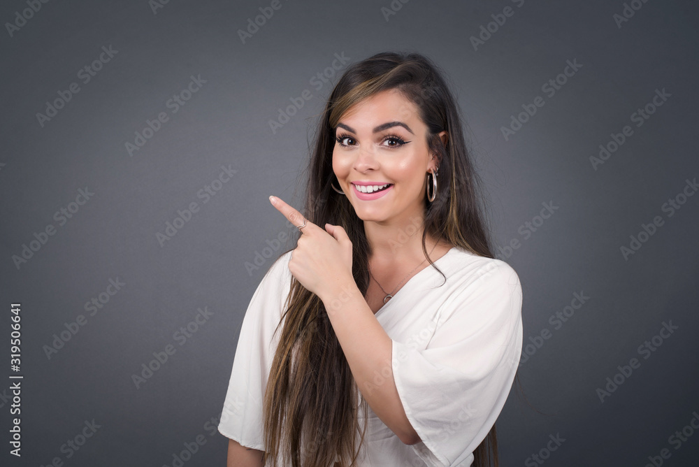 Look over there! Beautiful blonde caucasian woman pointing away and smiling to you while standing against grey background. Look over there! Beautiful blonde caucasian woman pointing away and smiling to you while standing against grey background.