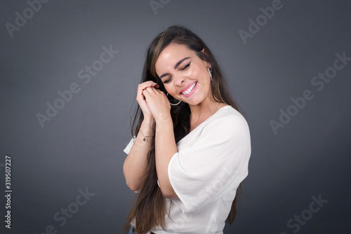 Dreamy young female with pleasant expression, closes eyes, keeps hands crossed near face, thinks about something pleasant, poses against gray background.