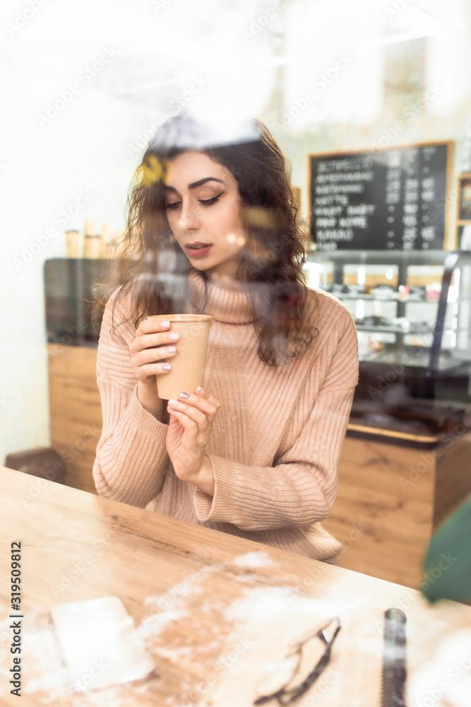 Girl holds a paper cup with coffee sitting at a wooden table in a coffee shop
