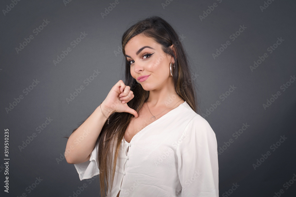 Fototapeta premium Closeup of cheerful beautiful European brunette young woman looks joyful, satisfied and confident, points at herself with thumb, isolated over gray studio background.
